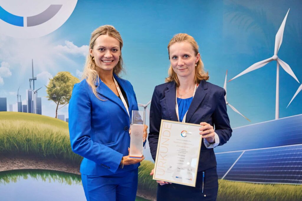 Linda Unser und Carola Stein halten beim Energy Transition Award 2024 eine Urkunde und einen Pokal vor einer Kulisse mit Windrädern und Solarpanelen
