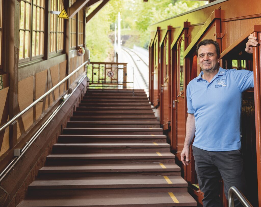 Martin Wenger, Wagenbegleiter bei den Heidelberger Bergbahnen, steht in blauer Polohemd am Eingang einer Bergbahnstation neben der Bahn