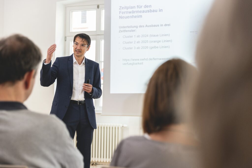 Thorsten Elfner hält einen kleinen Presenter in der Hand und gestikuliert mit der anderen. Hinter ihm wird eine Präsentation auf eine Leinwand projiziert.