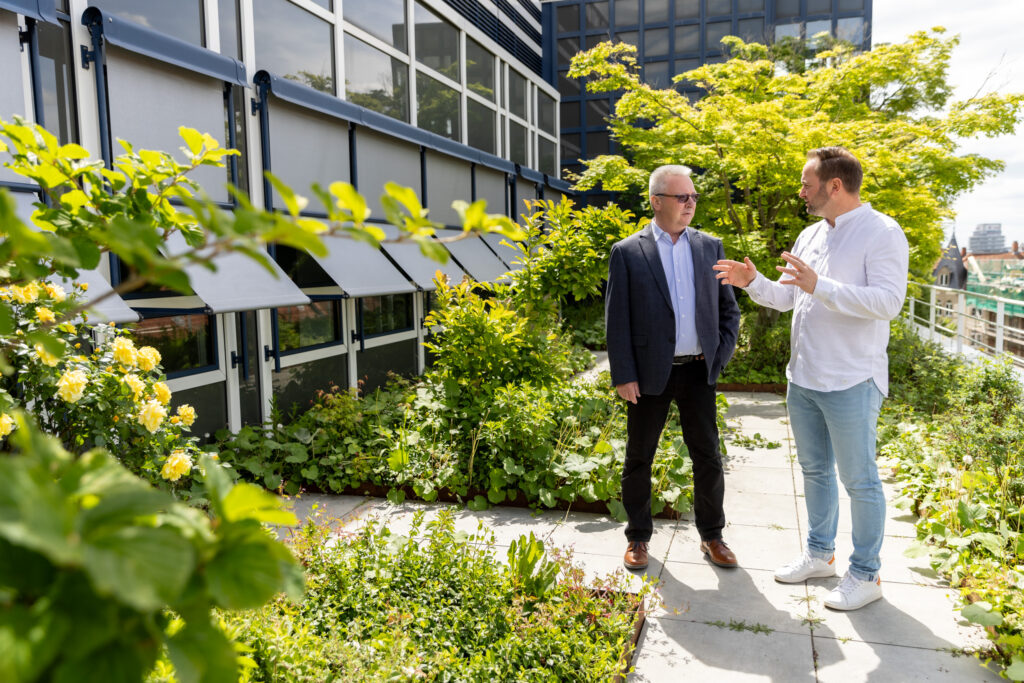 Peter Erb und Tobias Enders stehen auf einer Terrasse vor einem Bürogebäude, umgeben von grünen Pflanzen, im Gespräch