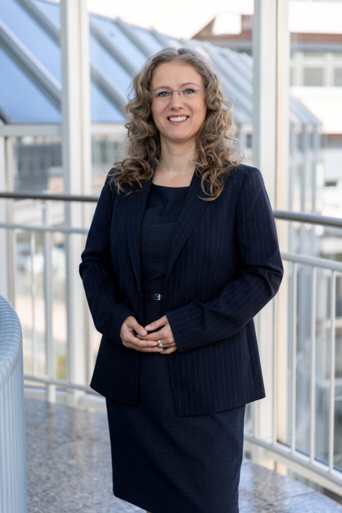 Meike Carstens steht in einem eleganten Anzug vor einem modernen Glasgebäude.