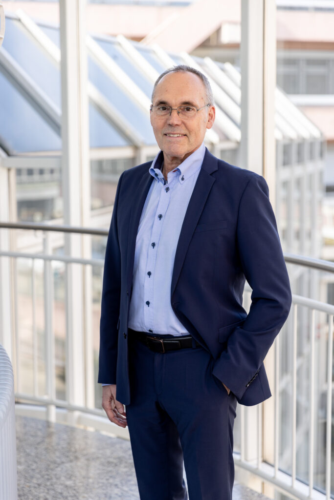 Rudolf Irmscher steht in einem Anzug vor einem modernen Glasgebäude.