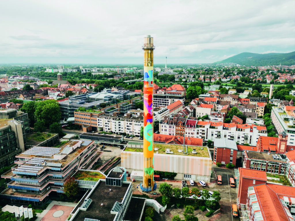Der Kaminturm des Heizwerks der Stadtwerke Heidelberg, bekannt als der heidelberg BLEISTIFT, mit bunter, abstrakter Bemalung inmitten der Stadt Heidelberg