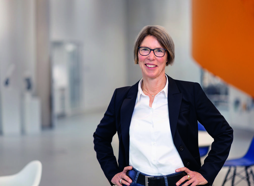 Karin Happes steht mit Händen in den Hüften in einem modernen, hellen Büro mit orangefarbenem Wandakzent, trägt weißen Hemd und dunklen Blazer