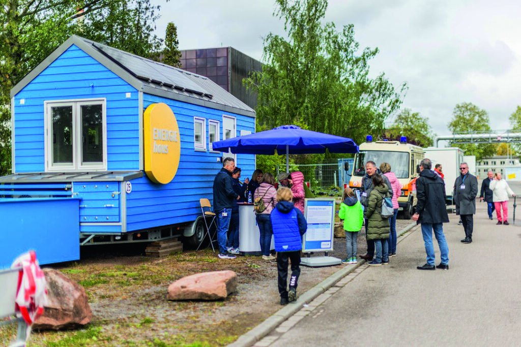 Menschen stehen an einem blauen Tiny House mit gelbem Schild 'ENERGIE HAUS' und einem blauen Sonnenschirm auf einem Gehweg im Freien