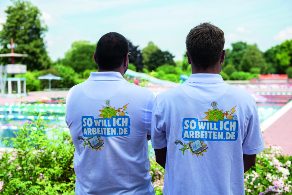 Zwei Personen in weißen Poloshirts mit Aufdruck 'SOWILLICHARBEITEN.DE' stehen vor einem Freibad mit Schwimmbecken und Rutschen, Beruf Fachangestellte für Bäderbetriebe