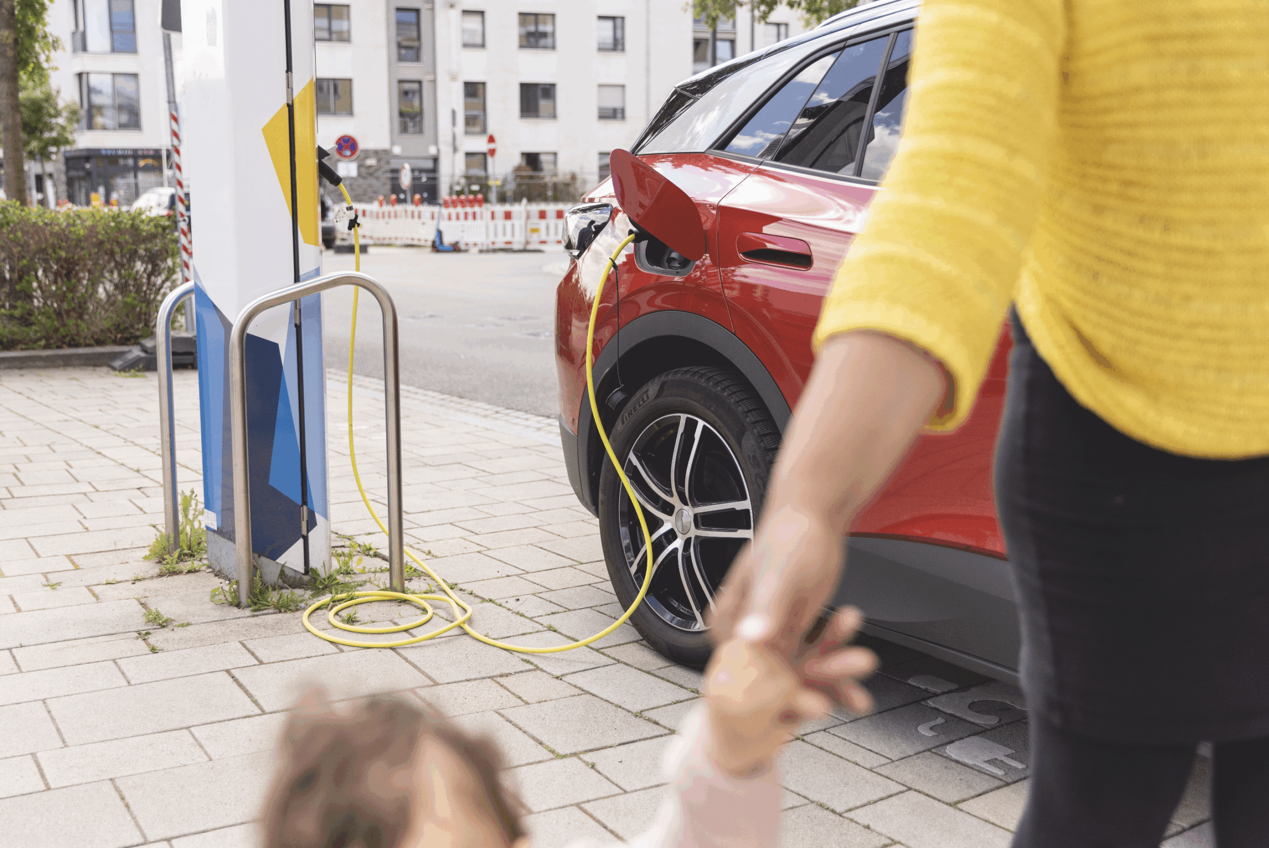 Amélie Martin hält die Hand ihrer Tochter, während ein rotes Elektroauto an einer Ladestation aufgeladen wird