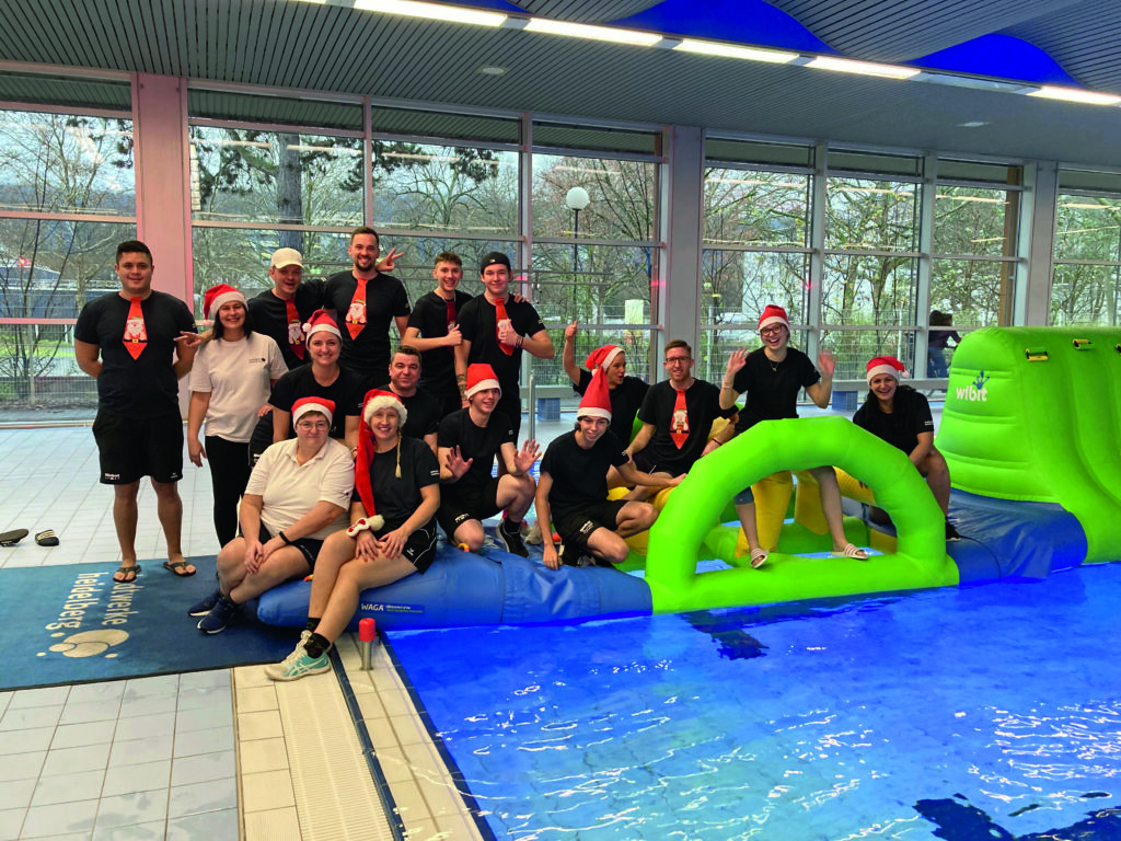 Personen in schwarzer Kleidung mit roten Weihnachtsmützen und roten Krawatten posiert an einem Hallenbad neben einem grünen und blauen aufblasbaren Wasserspielgerät