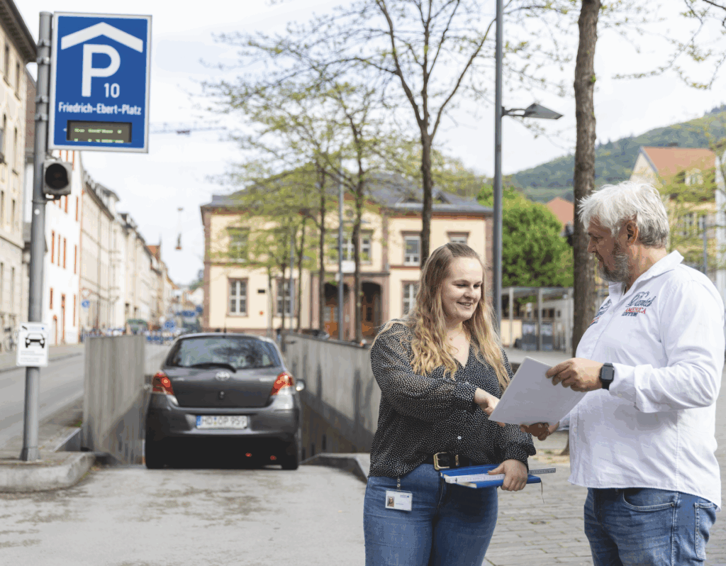 Thomas Buchwitz und seine Tochter Sarah Buchwitz aus dem Marketing-Team besprechen Dokumente auf einem Bürgersteig neben einem Parkhaus am Friedrich-Ebert-Platz