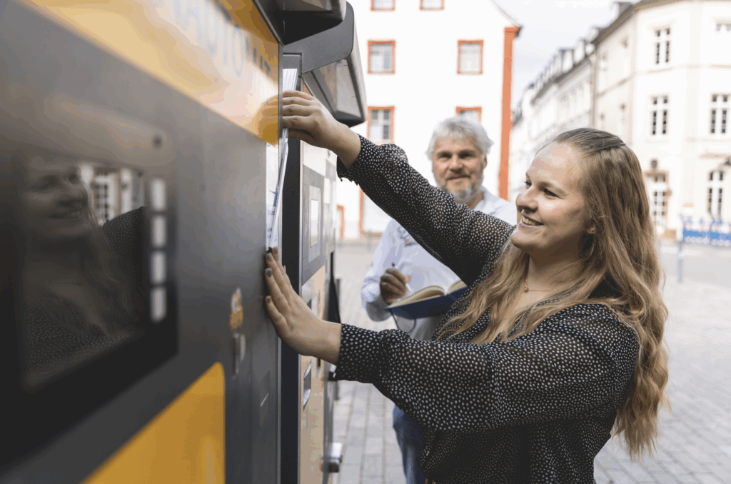 Sarah Buchwitz aus dem Marketing-Team bedient einen Parkautomaten auf der Straße, Thomas Buchwitz steht im Hintergrund mit Notizbuch