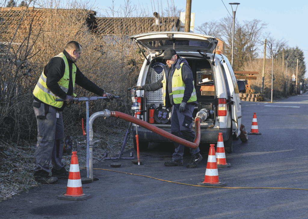 Zwei Arbeiter in gelben Warnwesten bedienen eine Maschine an einem weißen Transporter auf einer Straße, umgeben von Verkehrskegeln.