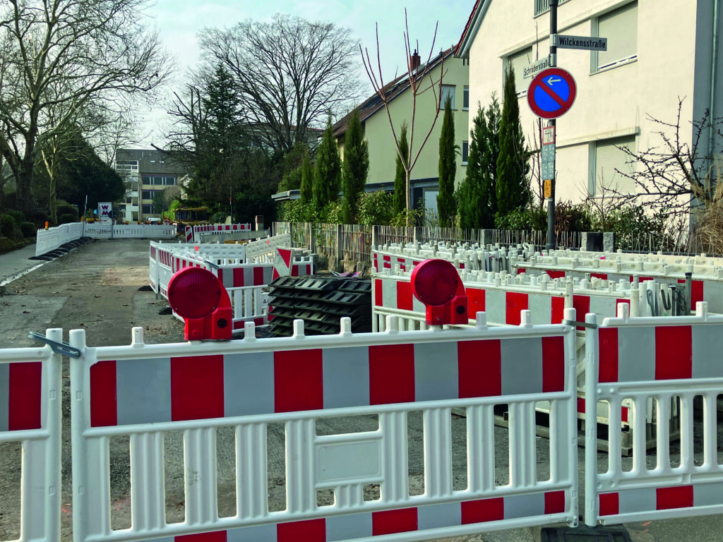 Straßensperrung mit weißen und roten Absperrungen und Warnleuchten in einer Wohnstraße mit Häusern und Bäumen