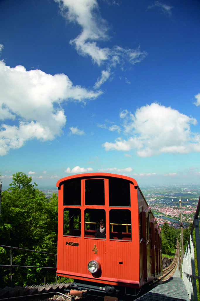 Rote Bergbahn fährt einen steilen Hang hinauf mit Blick auf Stadt und blauen Himmel mit Wolken