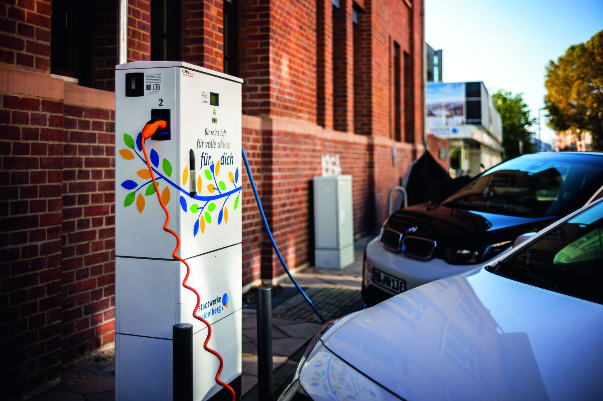 Elektroauto lädt an Ladestation mit Aufschrift 'Für unsere Welt, für volle Akkus, für dich' vor Backsteingebäude