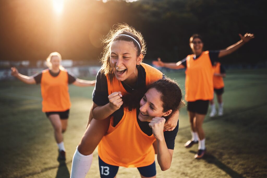 Junge Frauen im Fußballtraining mit orangefarbenen Trikots, eine Spielerin jubelt auf den Schultern einer anderen.