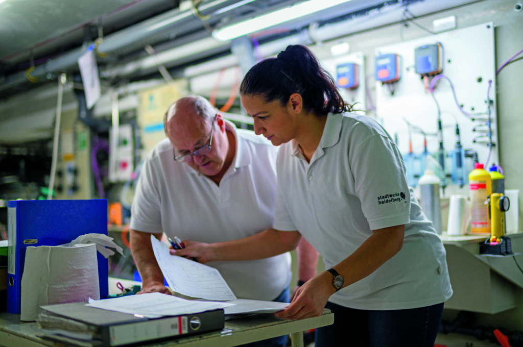 Zwei Personen in Arbeitskleidung besprechen Unterlagen in einem technischen Arbeitsumfeld zur Vorbereitung auf die Prüfung zum Nachhaltigen Wirtschaften