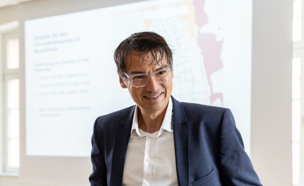 Thorsten Elfner steht in einem Anzug vor einem Projektorbildschirm und spricht mit Personen