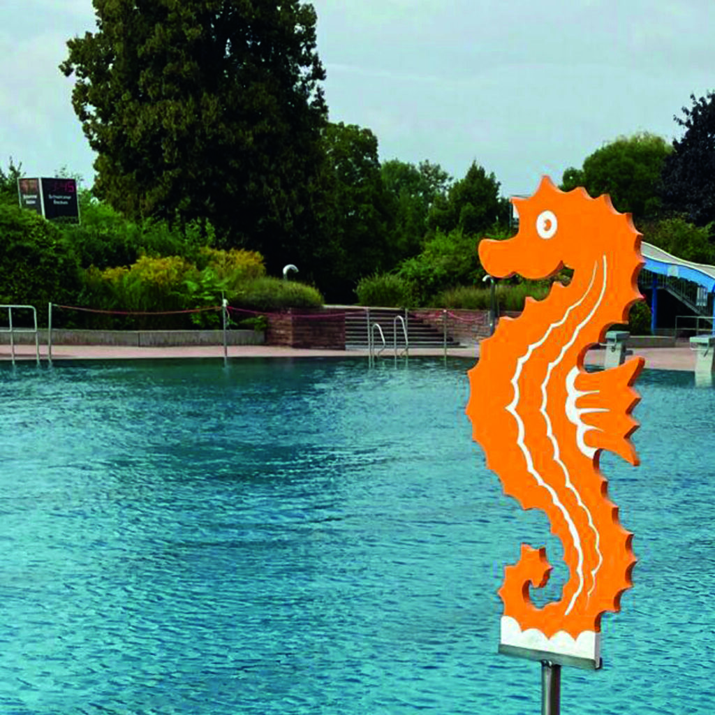 Orangenfarbenes Seepferdchen-Schild am Rand eines Schwimmbeckens mit Wasser und Bäumen im Hintergrund