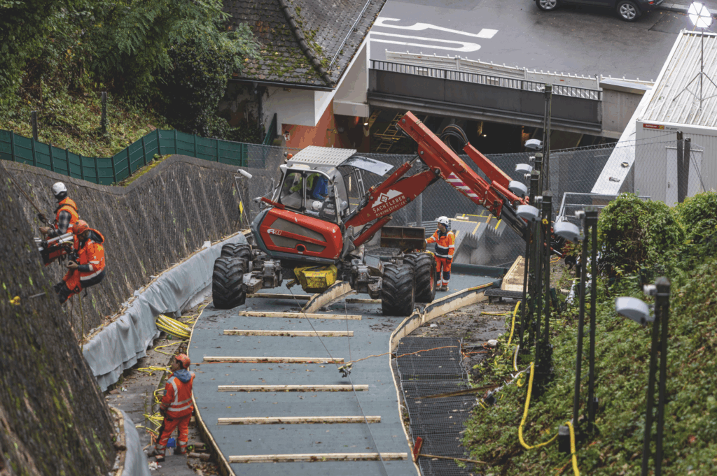 Arbeiter montieren ein Sicherungsnetz aus Stahl an der Stützmauer entlang der Bergbahnstrecke mit Hilfe eines roten Minibaggers