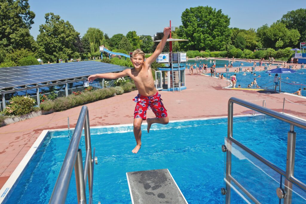 Junge in roten Badehosen springt vom Sprungbrett in ein Freibad mit mehreren Schwimmern und grünen Bäumen im Hintergrund