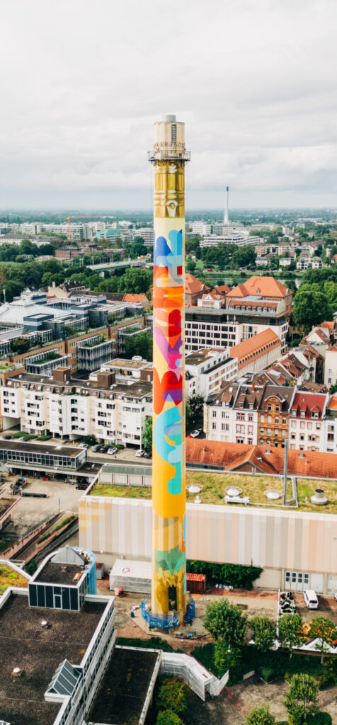 Der Kaminturm des Heizwerks der Stadtwerke Heidelberg, bekannt als der heidelberg BLEISTIFT, mit bunter, abstrakter Bemalung inmitten der Stadt Heidelberg
